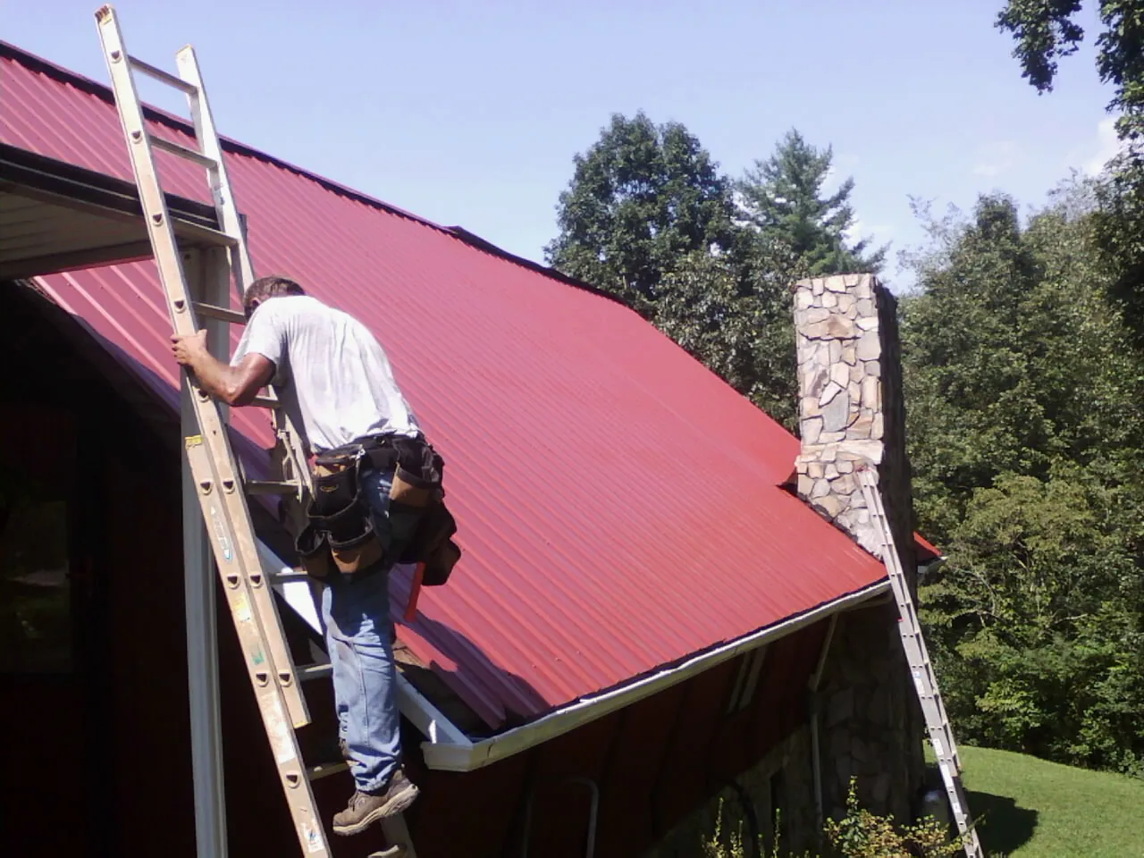 Roofer on a ladder beside a red metal roof and stone chimney — Soffit & Fascia Repair by Roofing Brothers in Bliss